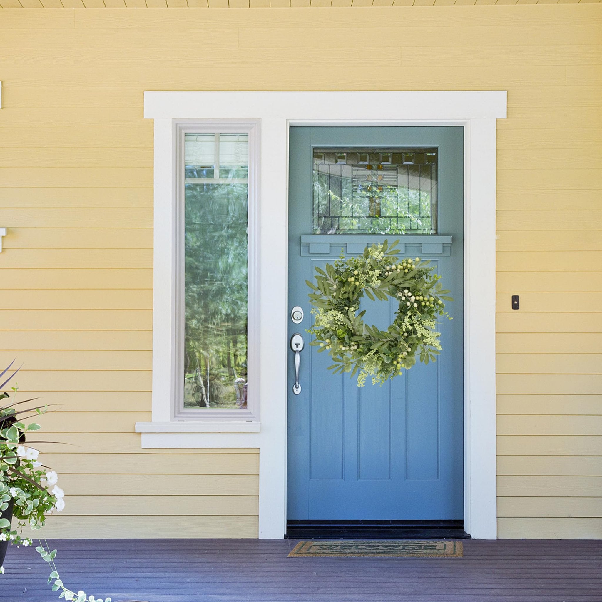 Eucalyptus and olive leaf wreath with white berries hanging on a blue front door with a window beside it.