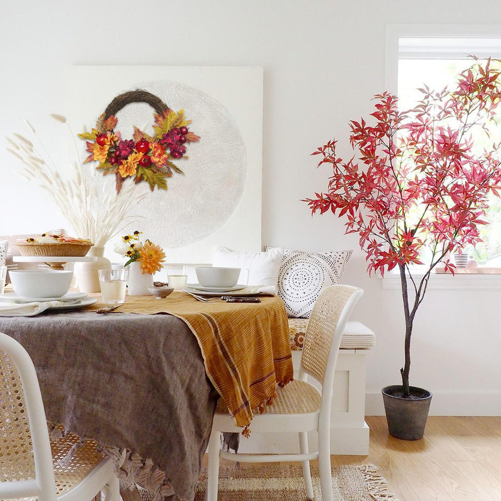 Fall wreath with apples, pomegranates, and maple leaves on a textured white circle backdrop above a dining table.