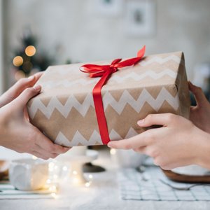Two hands exchanging a wrapped gift tied with a red ribbon on a table with festive lights.