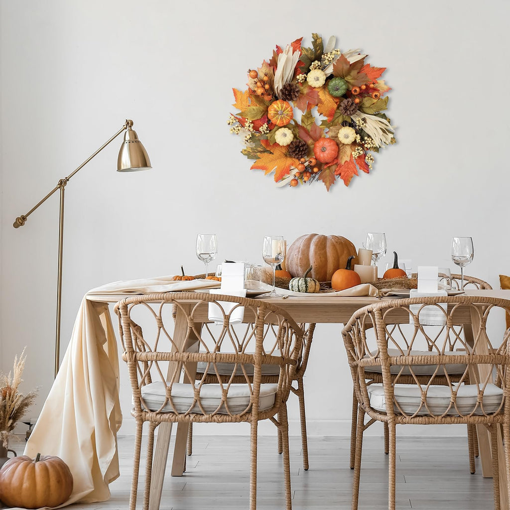 Fall wreath with pumpkins, maple leaves, berries, pine cones, and corn silk hanging on a wall above a dining table set for a meal.