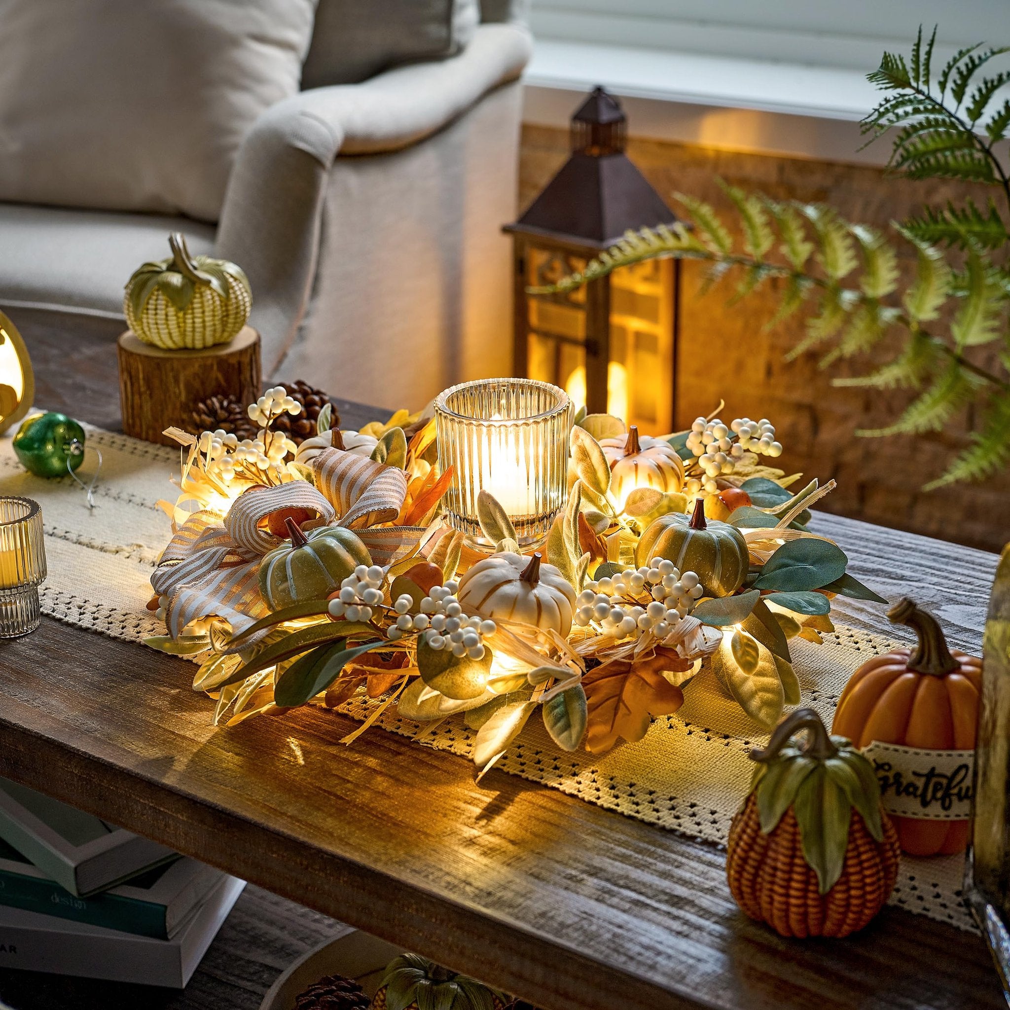 Lit fall wreath with pumpkins, berries, leaves, and burlap bow on a table with a candle and lantern.