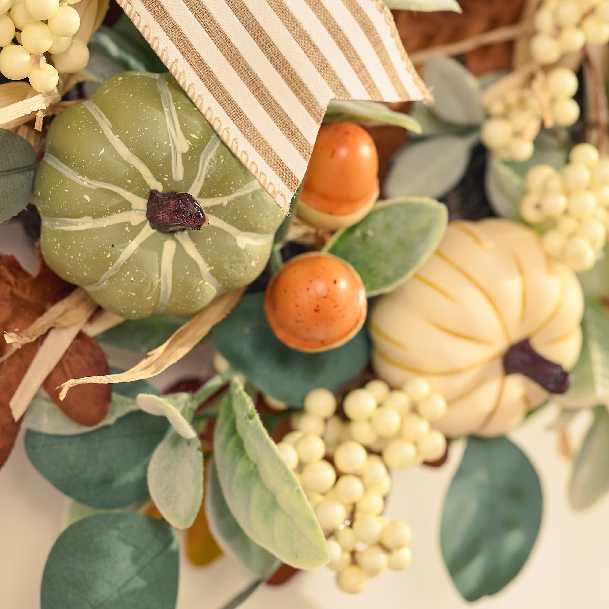 Close-up of a fall wreath with green and white pumpkins, orange berries, and a striped burlap bow.
