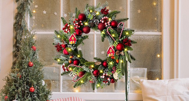 Lighted Christmas wreath with candy canes, pine cones, berries, red ornaments, and small house decorations hanging on a frosted window.