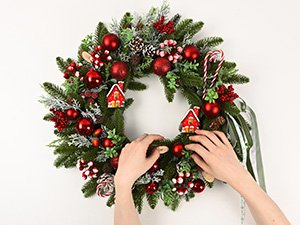 Hands decorating a 20-inch artificial Christmas wreath with candy canes, pine cones, berries, red ornaments, and small house ornaments.