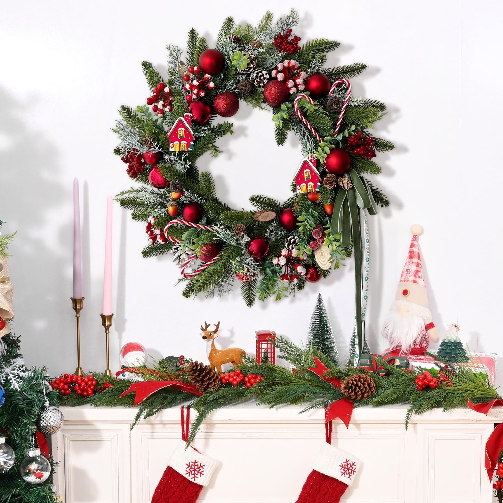 Festive Christmas wreath with red ornaments, pine cones, berries, candy canes, and small houses, displayed above a mantelpiece with garland and stockings.