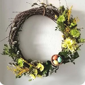 Decorated Easter wreath with pussy willow, green and yellow flowers, and a small nest with three pastel eggs, hanging on a white wall.
