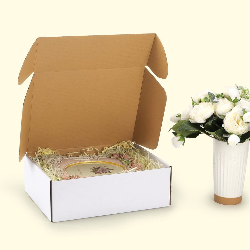Open white shipping box with decorative filler and a framed picture inside, next to a vase of white flowers.