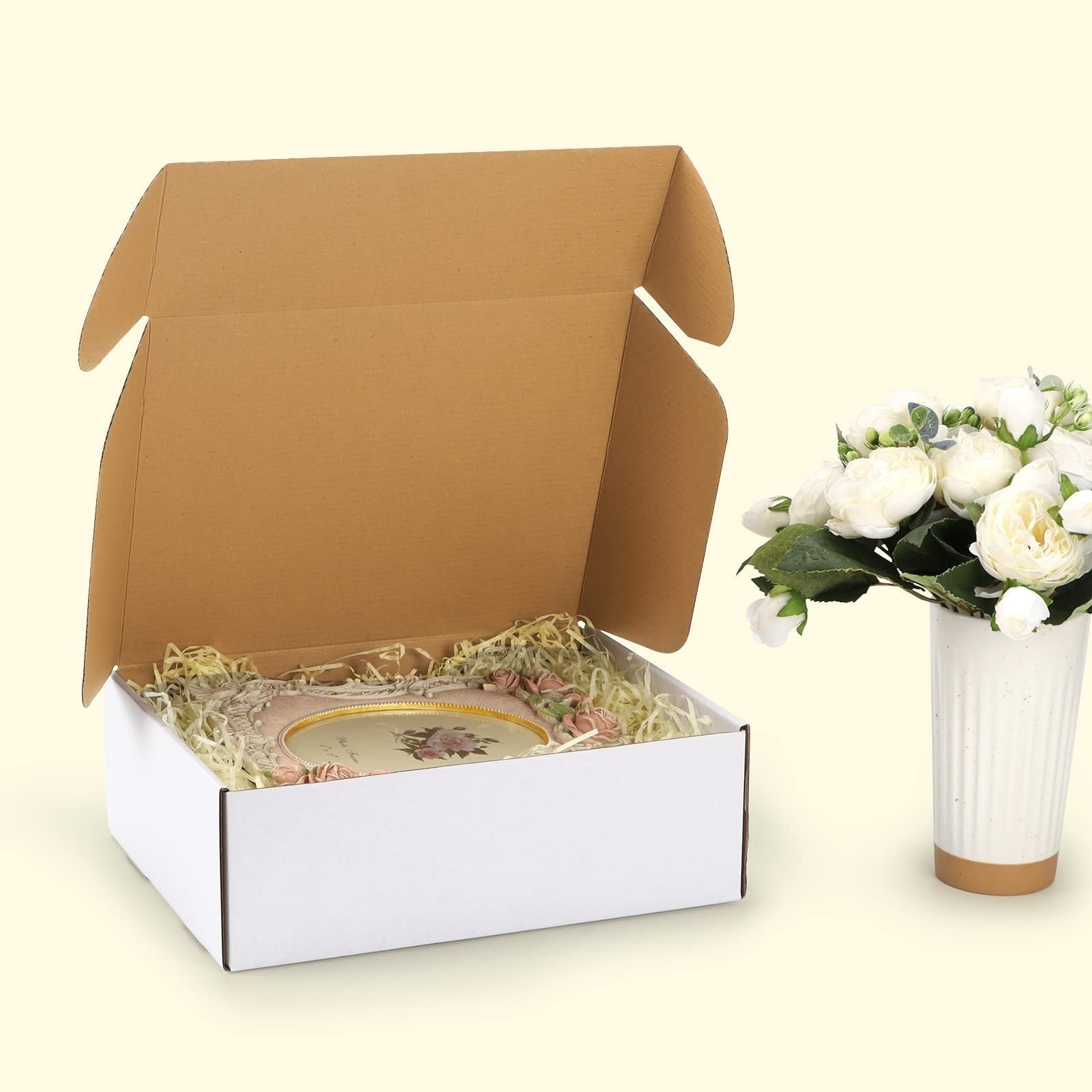 Open white shipping box with decorative filler and a framed picture inside, next to a vase of white flowers.
