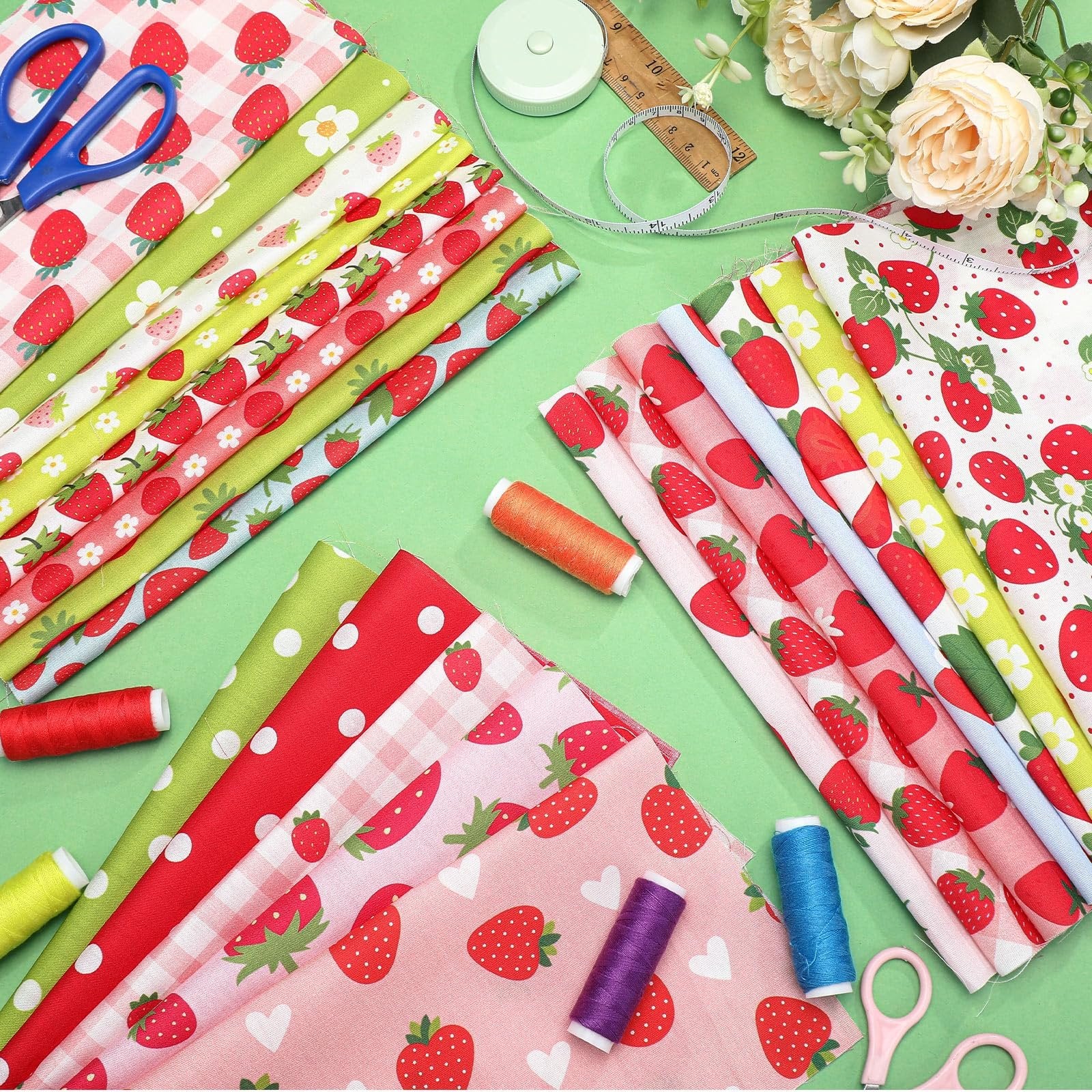 Assortment of strawberry-print fabric squares with spools of thread, scissors, and a measuring tape on a green background.