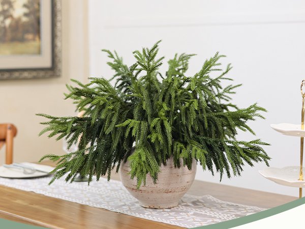Artificial Norfolk pine branches arranged in a vase on a table runner.