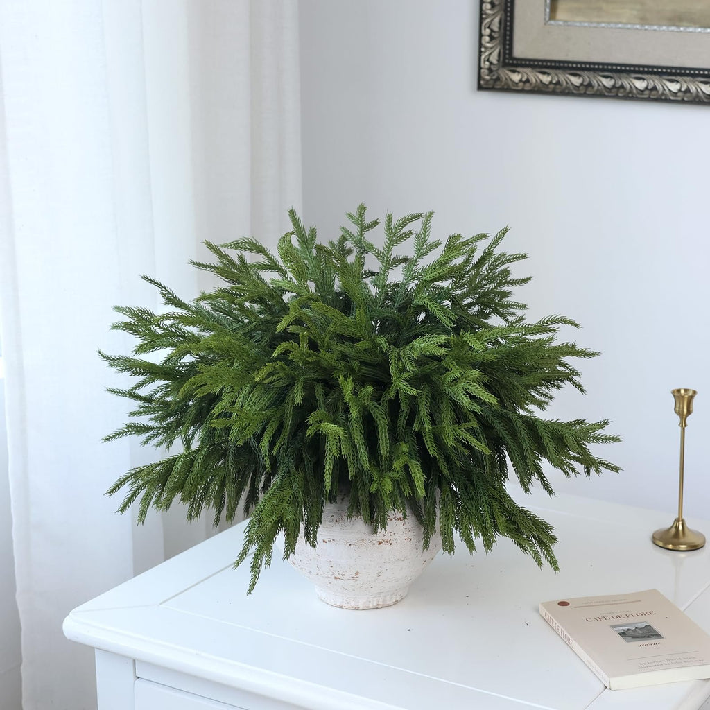 Artificial Norfolk pine branches in a textured white pot on a white table, with a book and candlestick nearby.