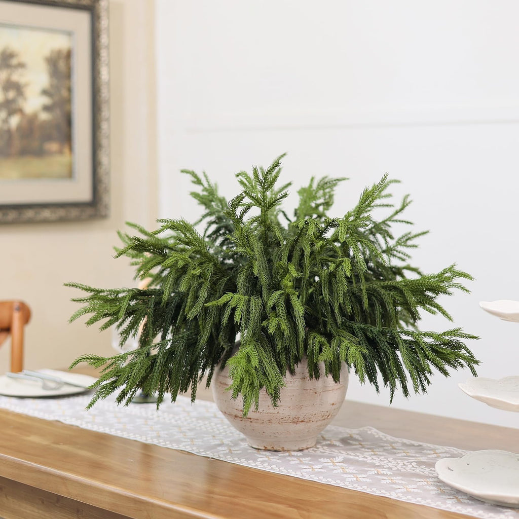 Artificial Norfolk pine branches arranged in a textured pot on a table runner with plates