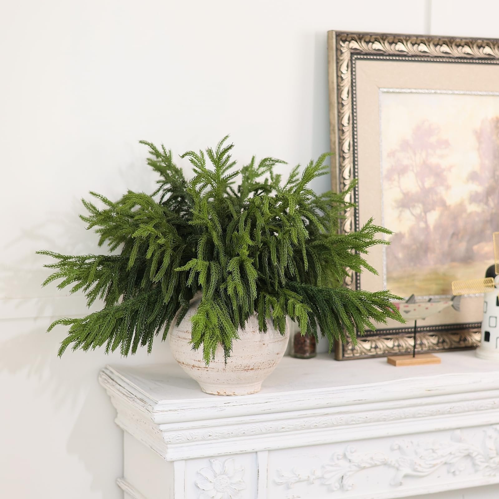 Artificial Norfolk pine branches arranged in a textured vase on a white mantelpiece, with a framed picture in the background.