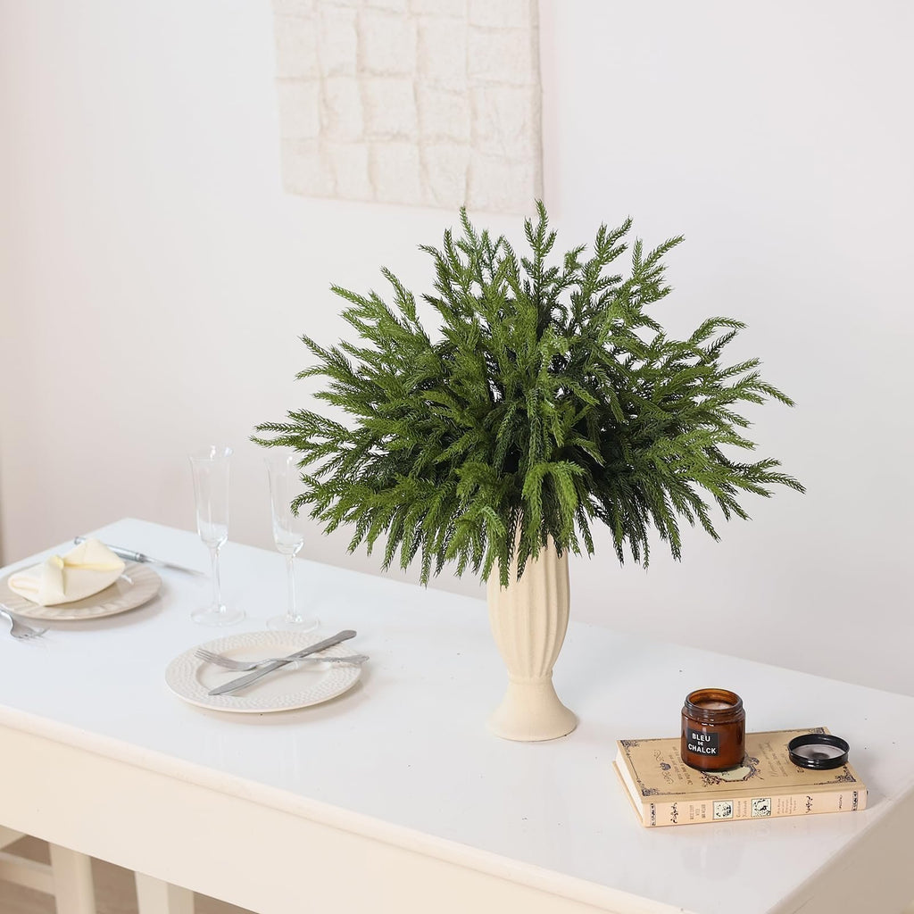 Artificial Norfolk pine branches in a vase on a table with place settings and a candle on a book