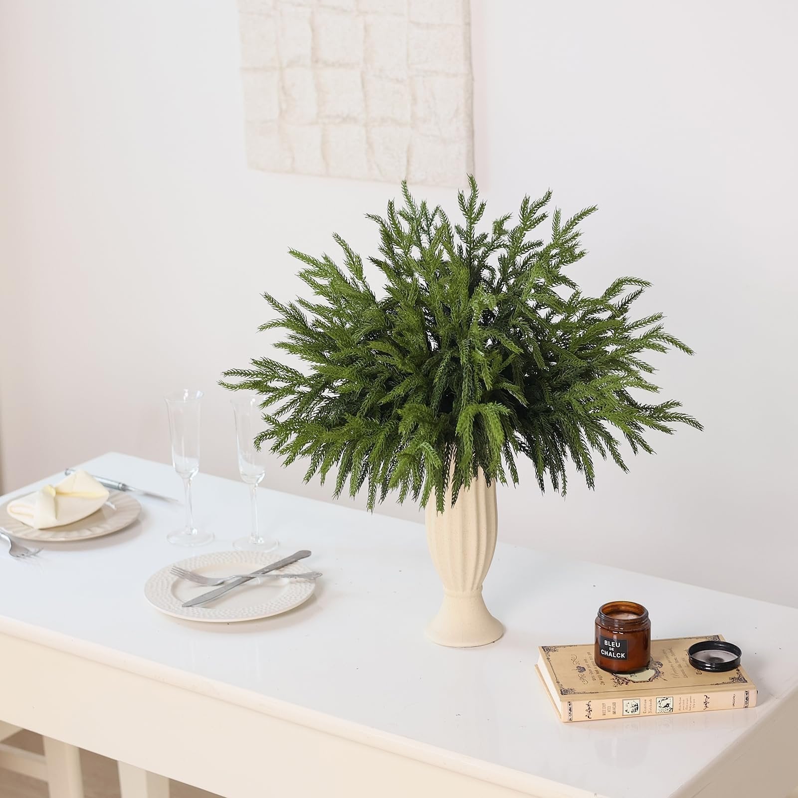 Artificial Norfolk pine branches in a vase on a table with place settings and a candle on a book