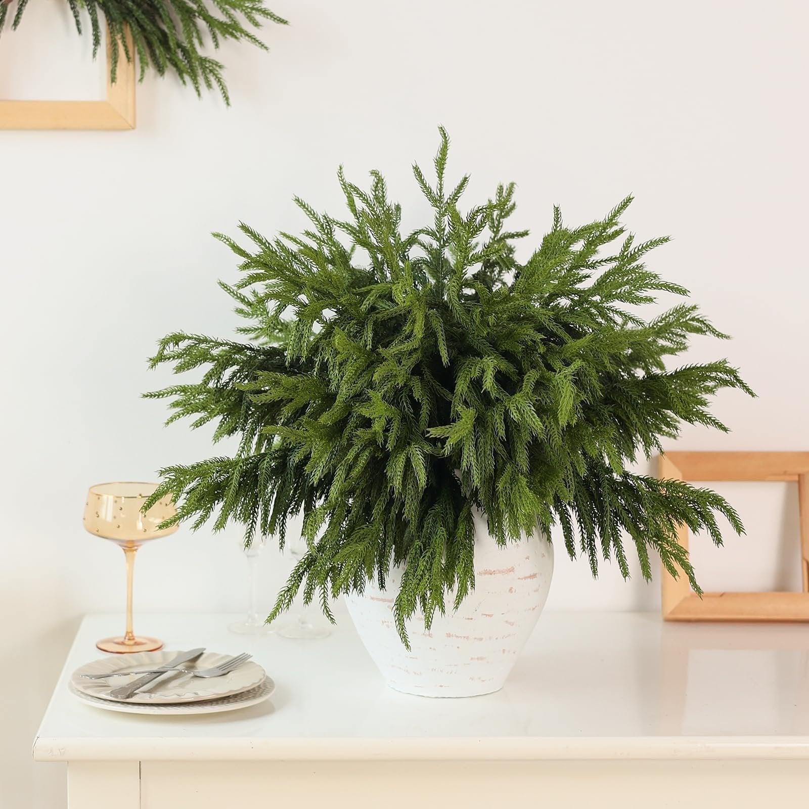 Artificial Norfolk pine branches arranged in a white vase on a table with place settings and decorative frames.