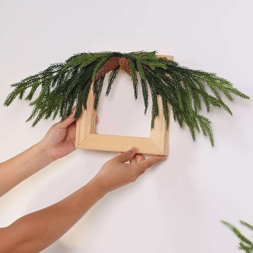 Hands holding a wooden frame decorated with artificial pine branches and pinecones