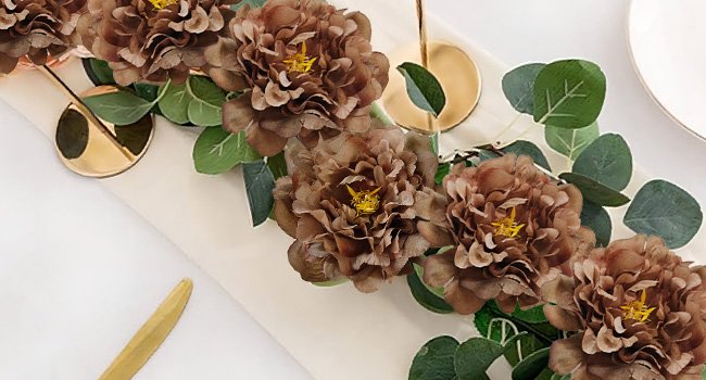 Brown artificial peony flowers with eucalyptus leaves arranged on a table runner with gold accents.