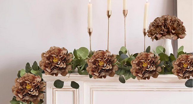 Brown artificial peony flowers and eucalyptus garland draped over a mantelpiece with candlesticks