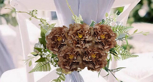 Five brown artificial peony flower heads with yellow centers, arranged on a white chair with greenery.