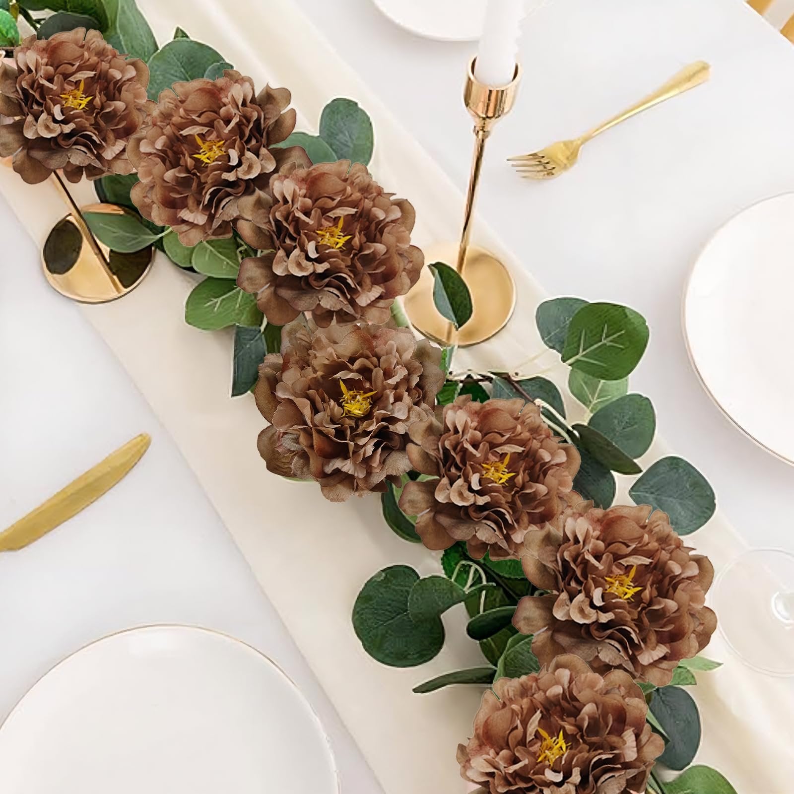 Brown artificial peony flowers and eucalyptus leaves arranged on a table runner with place settings.