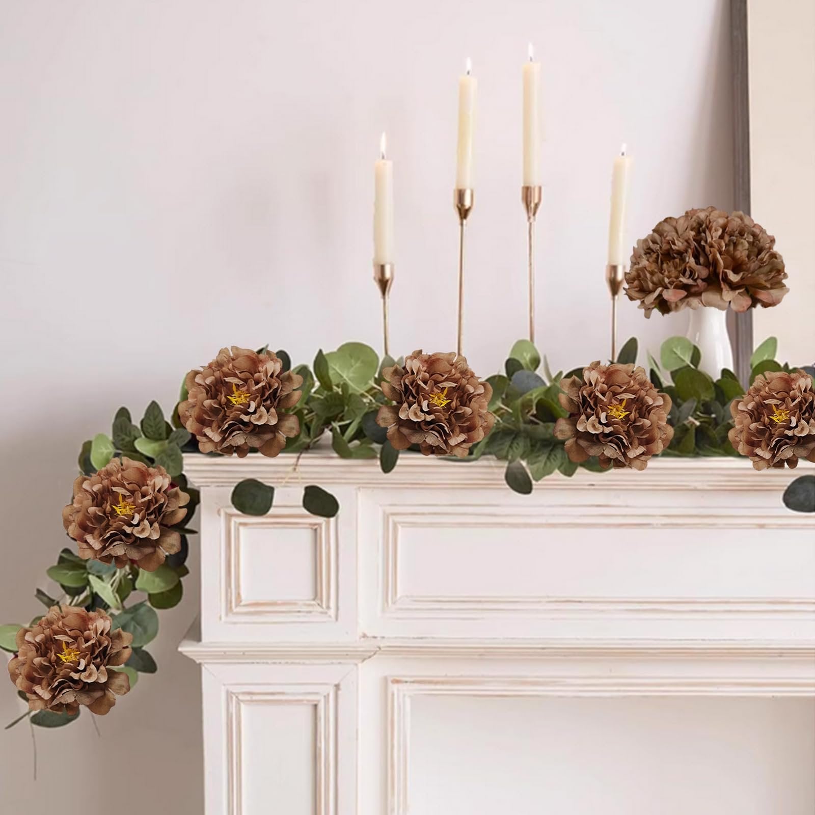 Brown artificial peony flowers and eucalyptus garland draped over a white mantelpiece with candlesticks