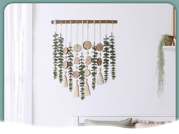 Boho wall hanging with dried eucalyptus branches, wood slices, beads, and tassels on a white wall.
