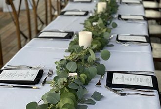 Table setting with white tablecloth, black napkins, menus, cutlery, and a eucalyptus garland centerpiece with candles.