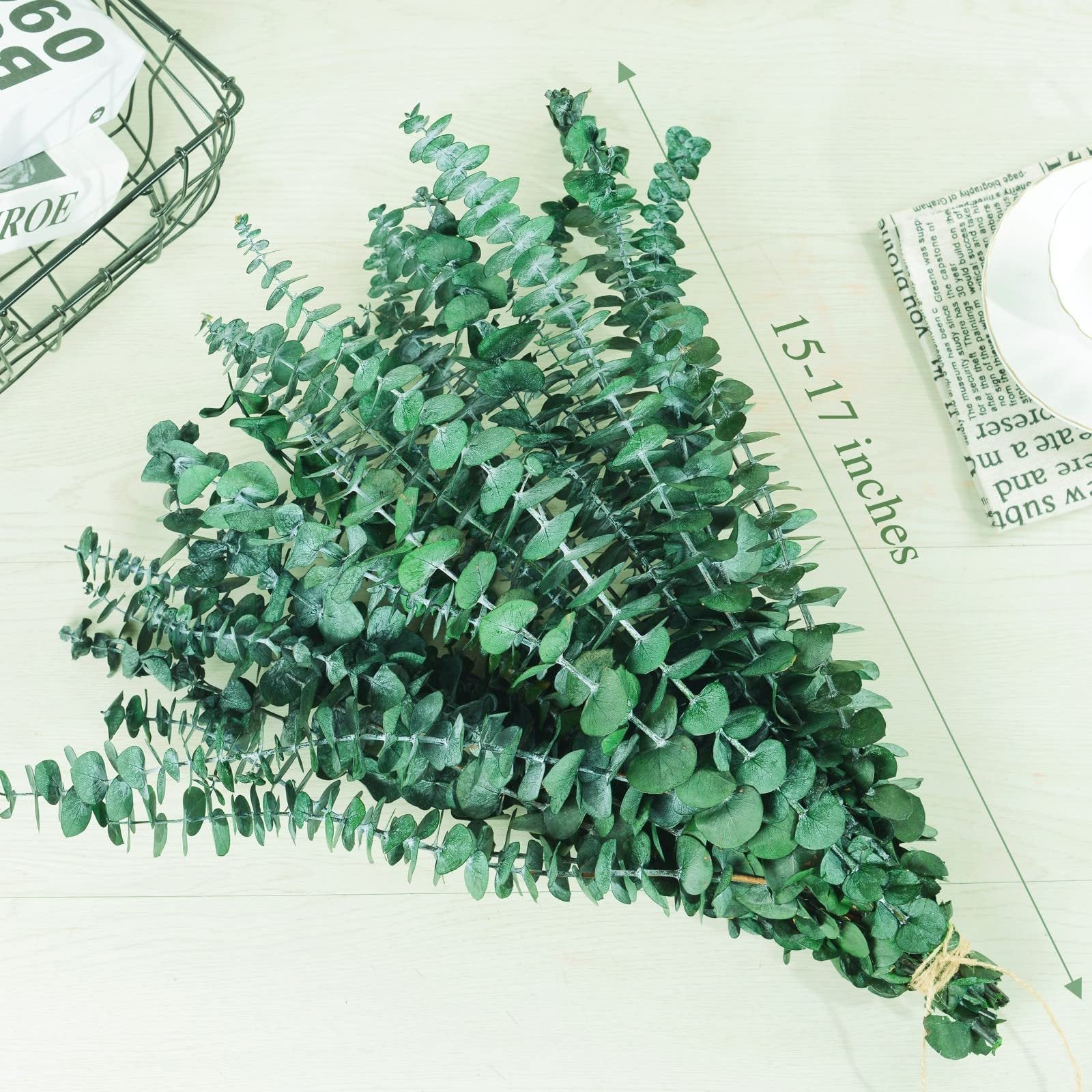 Bundle of 20 preserved dried eucalyptus stems, 15-17 inches long, on a light wood surface with a basket and newspaper in the background.