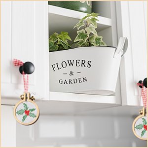 Mini wood embroidery hoops with holly pattern hanging on cabinet knobs, next to a planter with plants.