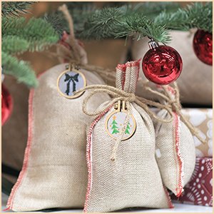 Mini wood embroidery hoops with festive designs used as gift tags on burlap bags under a Christmas tree.