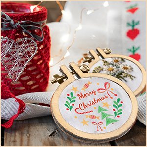 Two mini wood embroidery hoops with Christmas designs, one reading 'Merry Christmas', on a wooden surface with festive lights.