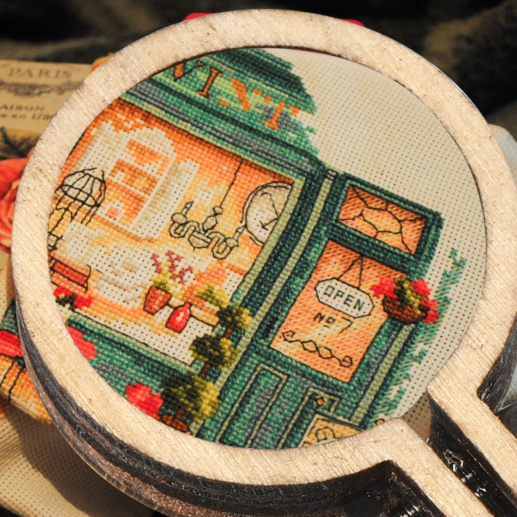 Close-up of a mini wood embroidery hoop with a cross-stitch design of a shop facade, showing an 'OPEN NO 7' sign.