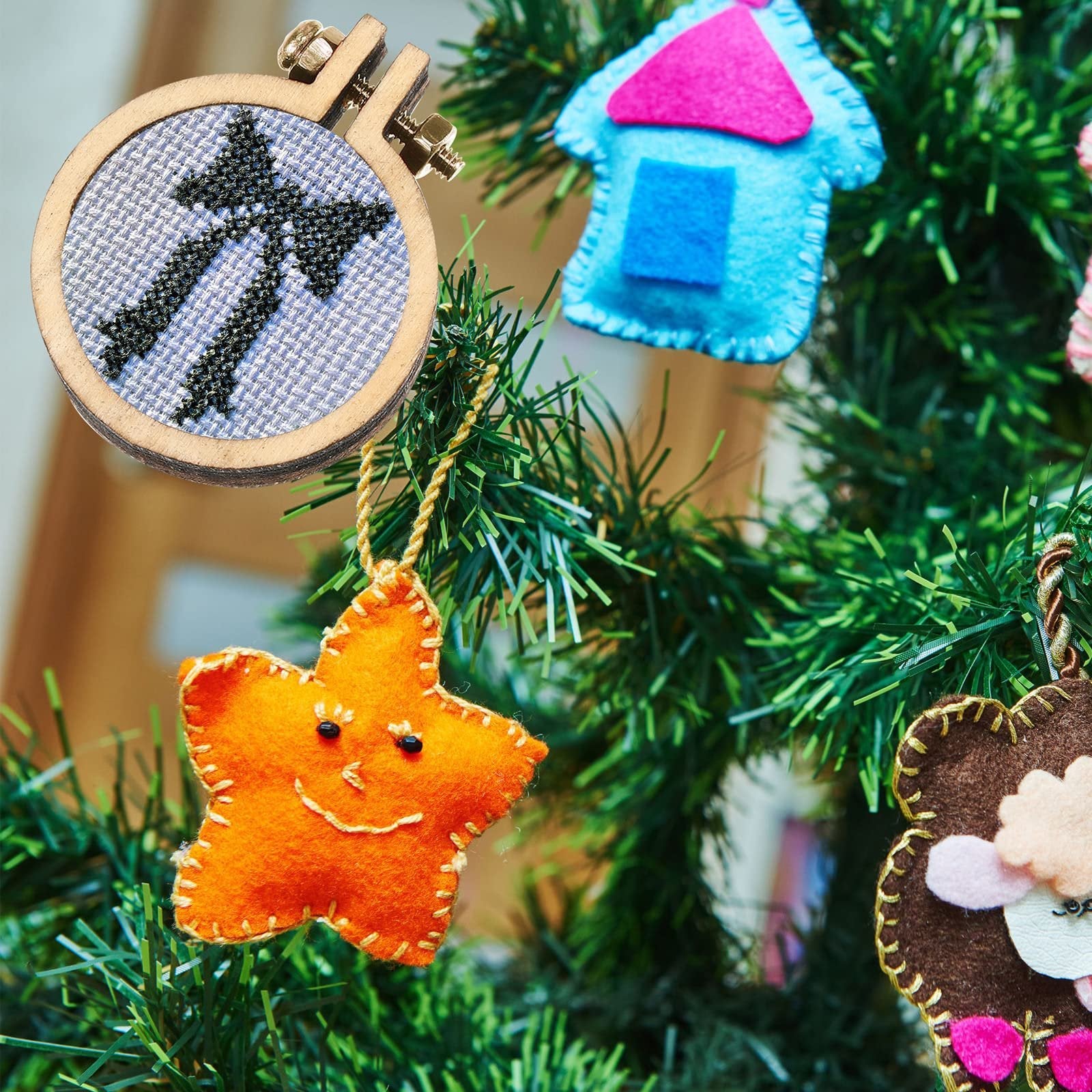 Mini wood embroidery hoop with black cross-stitch design hanging on a Christmas tree next to felt ornaments.