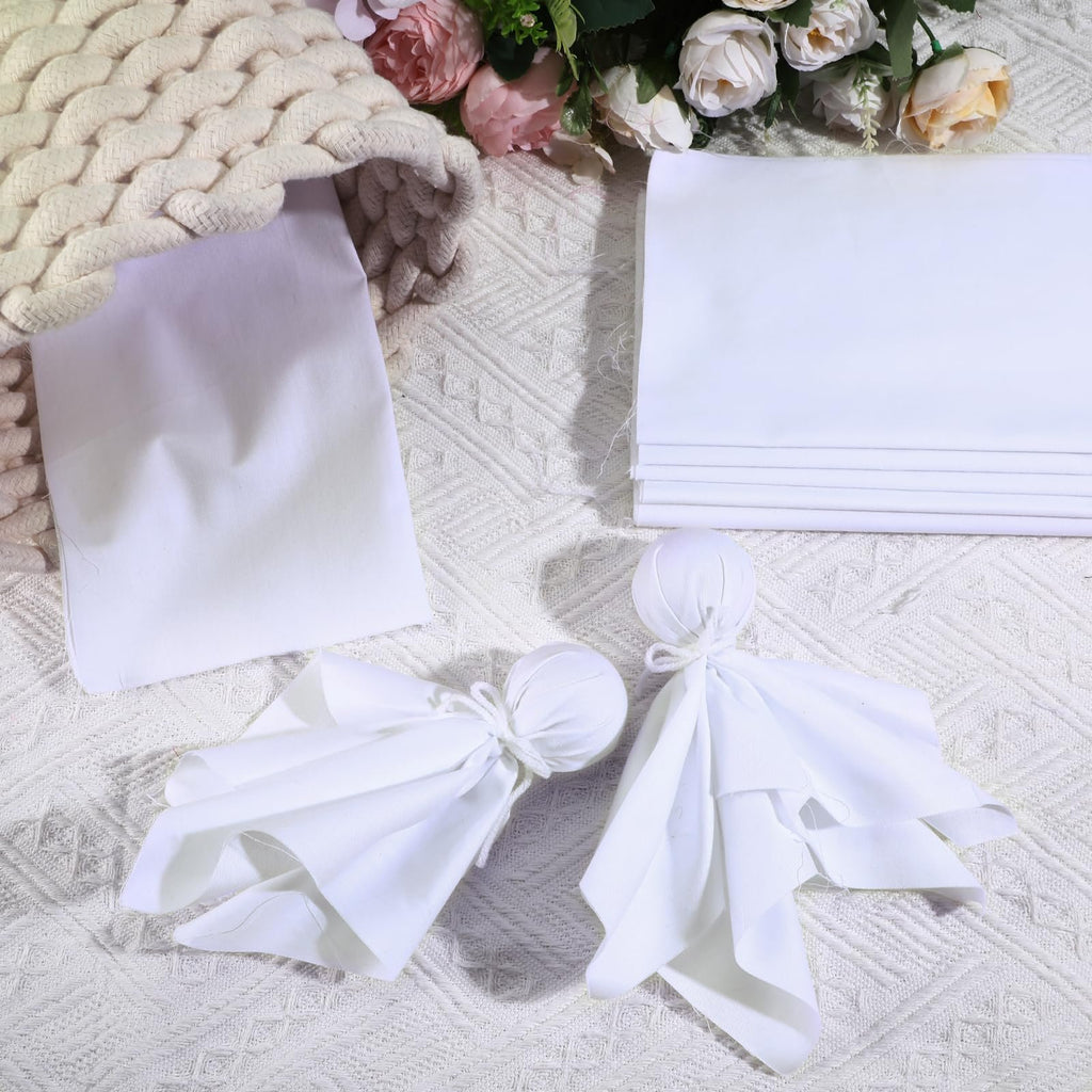 White cotton fabric squares and two fabric dolls arranged with a knitted basket and flowers on a textured white surface.