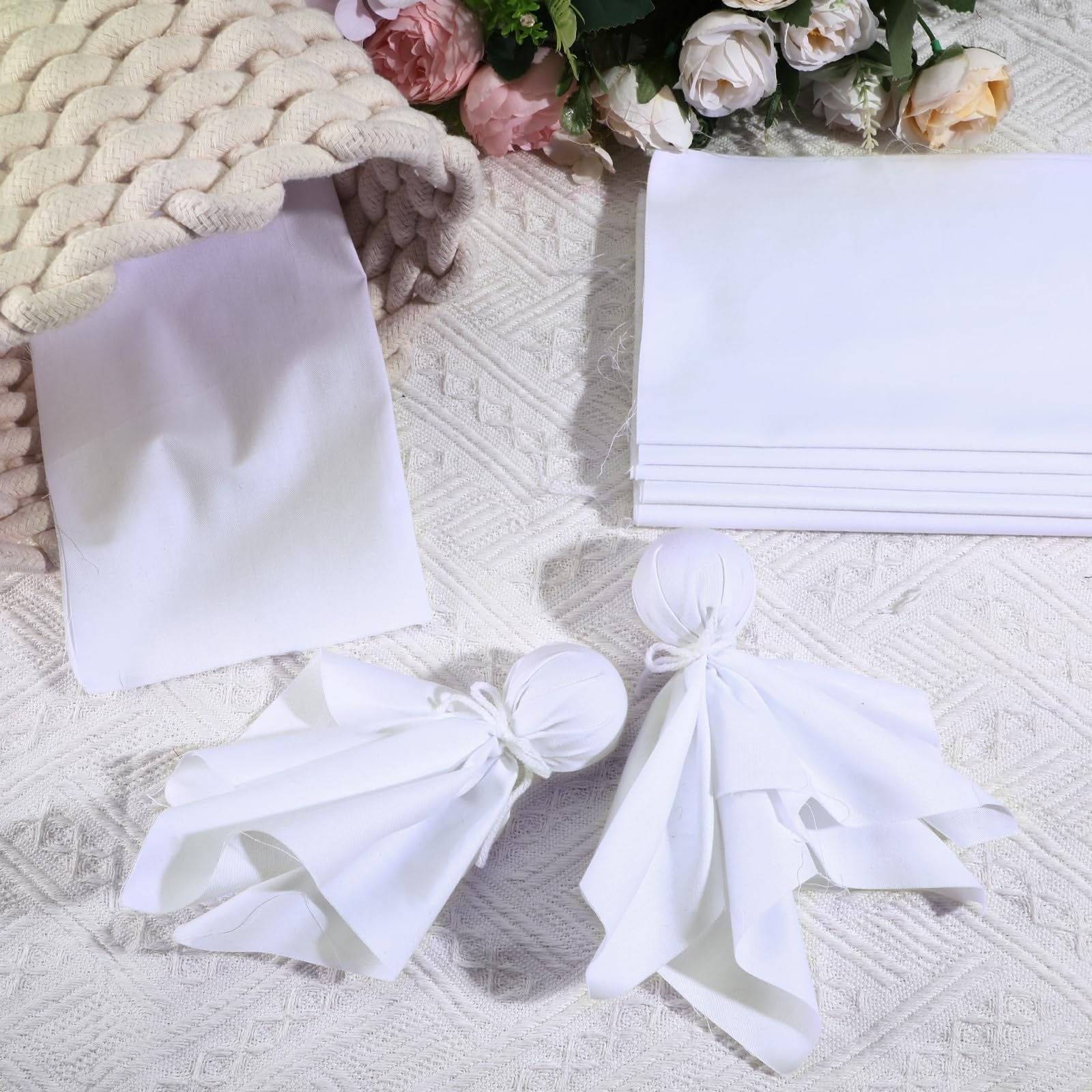 White cotton fabric squares and two fabric dolls arranged with a knitted basket and flowers on a textured white surface.