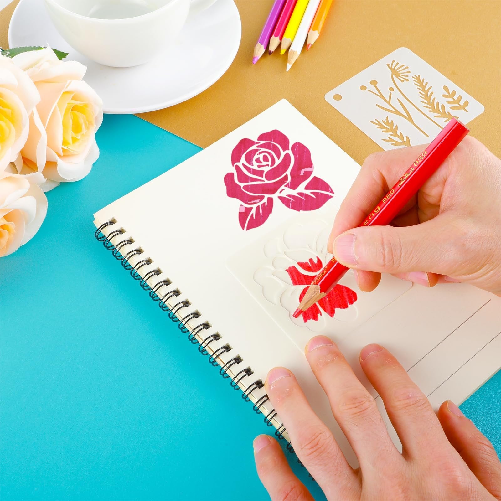 Hands using a flower stencil and red pencil to draw on a notebook, with colored pencils and roses nearby.