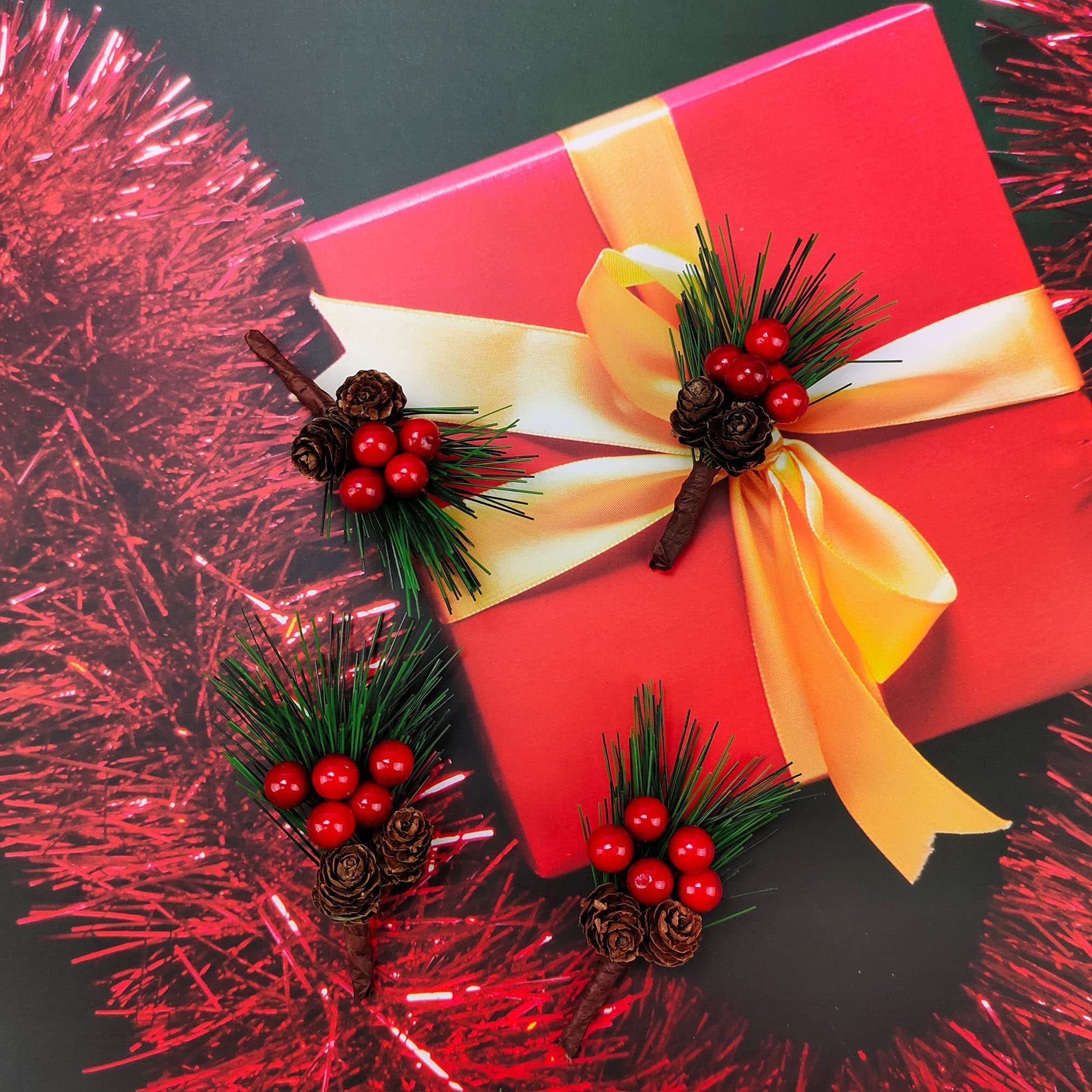 Christmas pine picks with red berries and pinecones arranged around a red gift box with a yellow ribbon.
