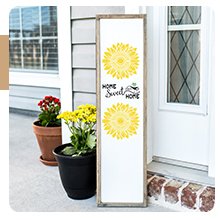 Tall wooden sign with 'HOME SWEET HOME' and two sunflower designs, placed on a porch with potted plants.
