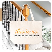Canvas tote bag with orange and black text reading 'this is us our life. our story. our home' hanging on a wooden banister.