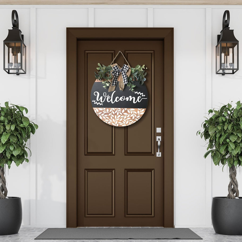 Round welcome sign with greenery and buffalo plaid bow hanging on a brown door, flanked by potted plants and wall lanterns.