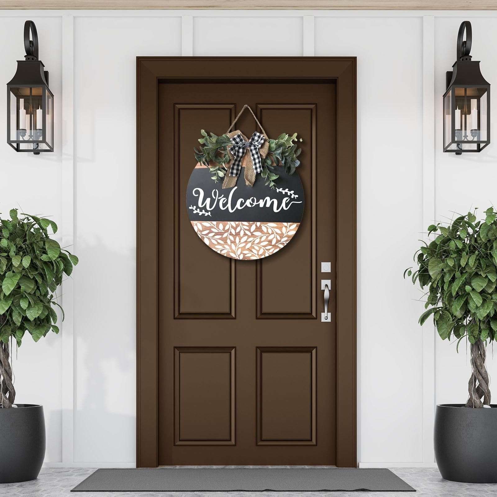 Round welcome sign with greenery and buffalo plaid bow hanging on a brown door, flanked by potted plants and wall lanterns.