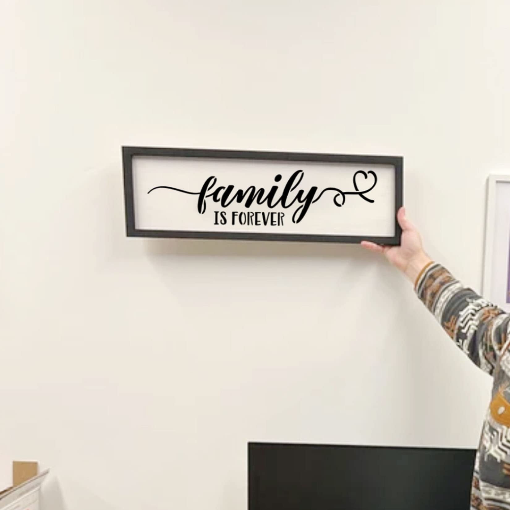 Framed farmhouse sign with 'family is forever' text and a heart, held by a person against a white wall.