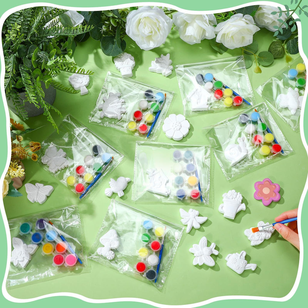 Ceramic painting kit with plaster figurines, paints, and brushes laid out on a green surface with floral decorations.
