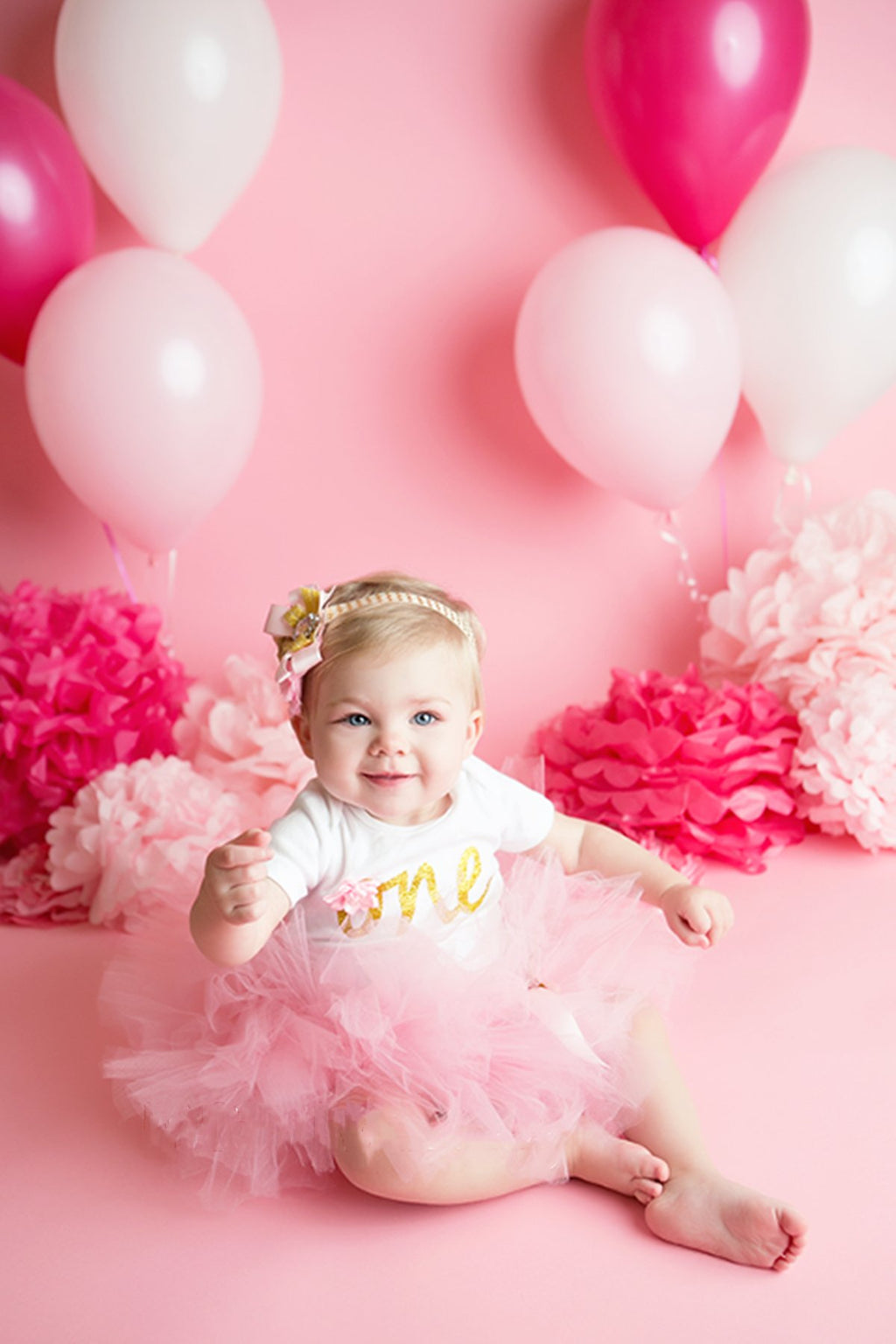 Baby girl in pink tutu and white shirt with 'one' in gold, sitting among pink balloons and tissue paper pom poms.