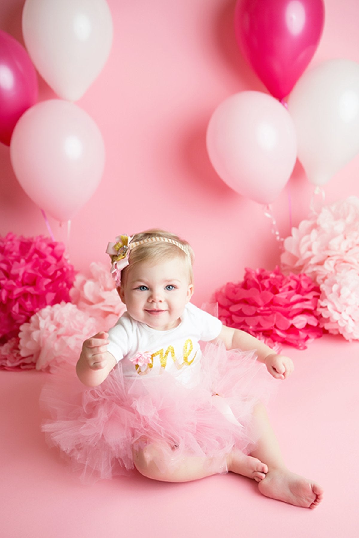 Baby girl in pink tutu and white shirt with 'one' in gold, sitting among pink balloons and tissue paper pom poms.