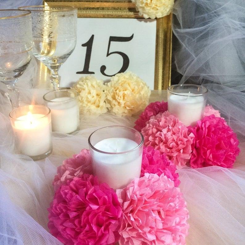 Table setting with pink and yellow tissue paper pom poms surrounding candles, with wine glasses and a table number '15' in the background.