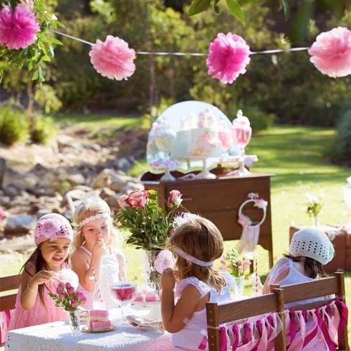 Four young girls in pink dresses at a garden party table with a cake and flowers, pink tissue paper pom poms strung overhead.