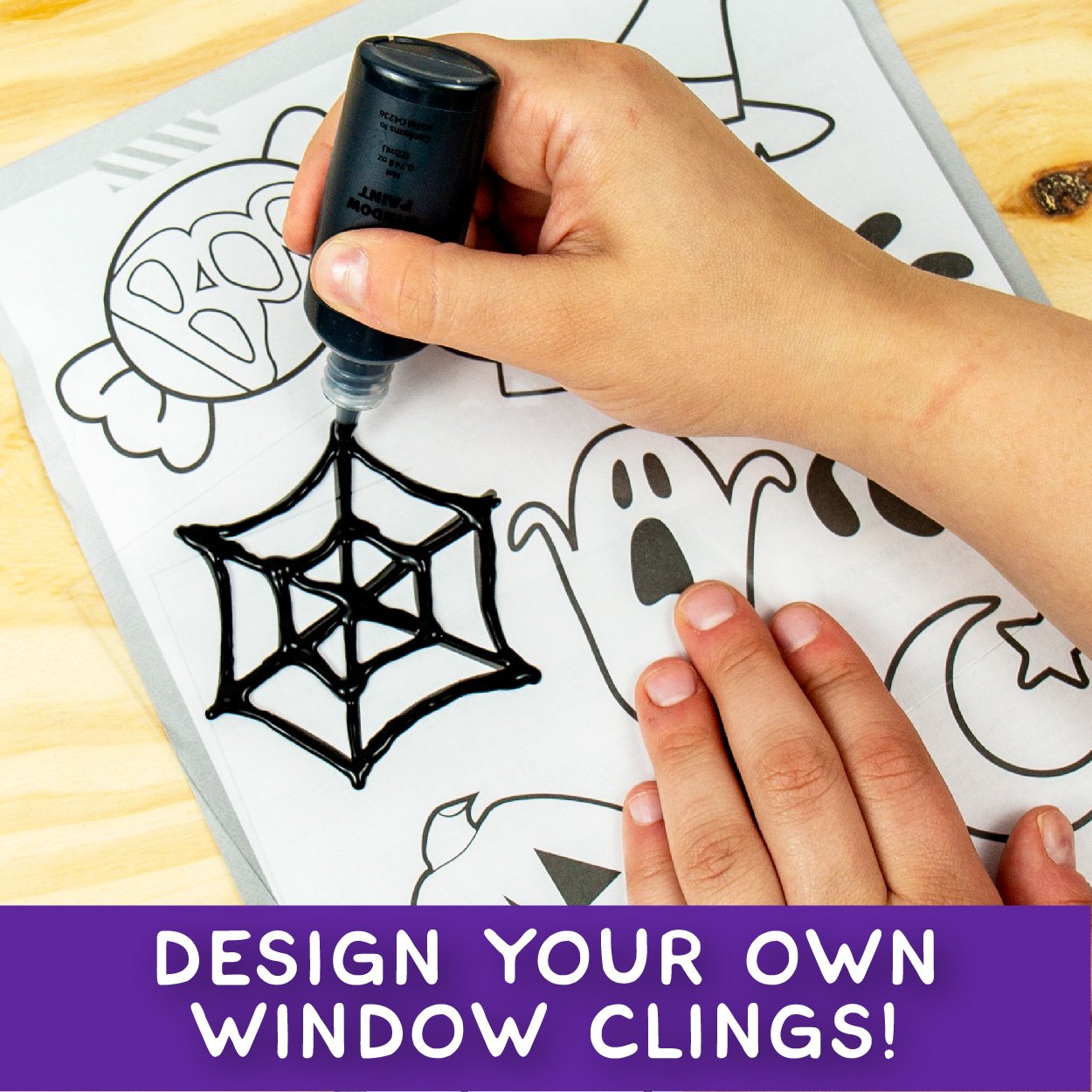 Child's hands using black window paint to trace a spider web design on a Halloween-themed template.