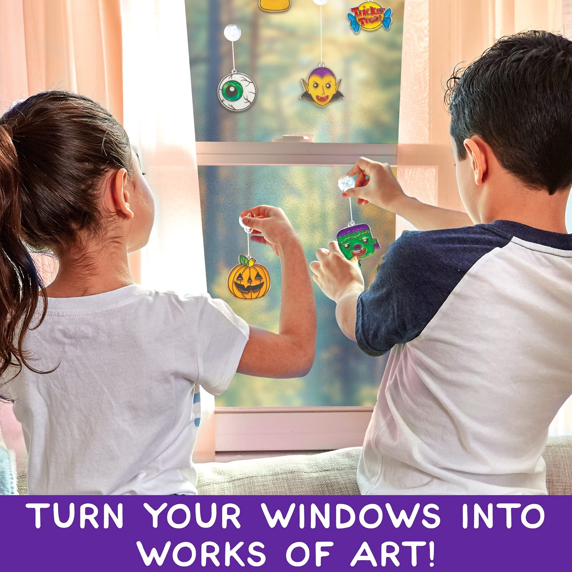 Two children hang Halloween-themed window art decorations on a window.